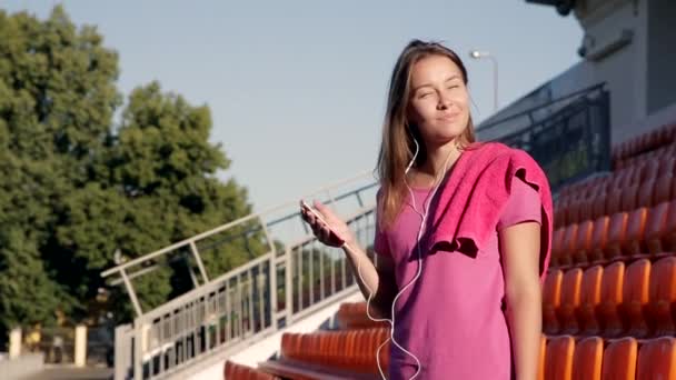 Joven chica atlética atractiva escuchando música en el estadio. Ella está vestida con una camiseta rosa. El sol de primavera ilumina su cara
. - Metraje, vídeo