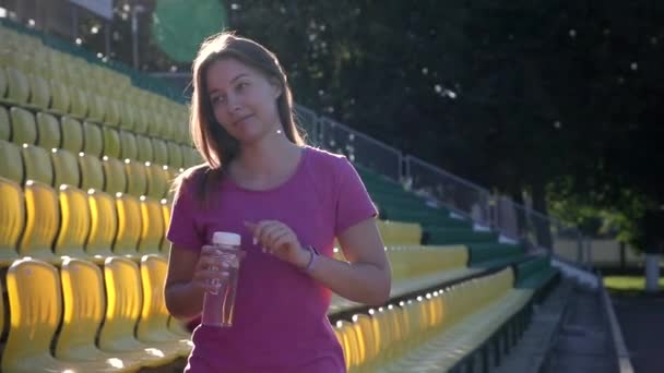 Young attractive woman is drinking water after sport training. She is dressing in a pink t-shirt. The morning spring sun warms her. - Imágenes, Vídeo