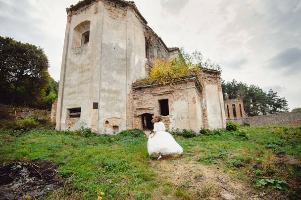 Het meisje in een witte jurk loopt op de achtergrond van het oude verlaten landgoed, op het dak waarvan struiken en bomen groeien. De ruïnes van het oude kasteel. - Foto, afbeelding
