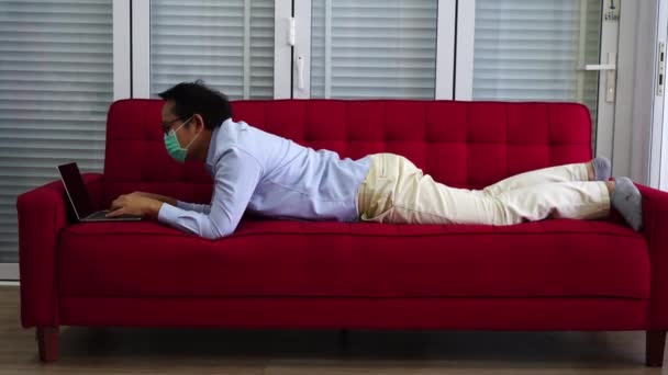 アジアの男性労働者は、彼がコロナ感染やcovidを防ぐために自宅に拘束された間、彼は赤いソファの上にラップトップで自宅から働くことに疲れていたので、眠っていました。 - 映像、動画