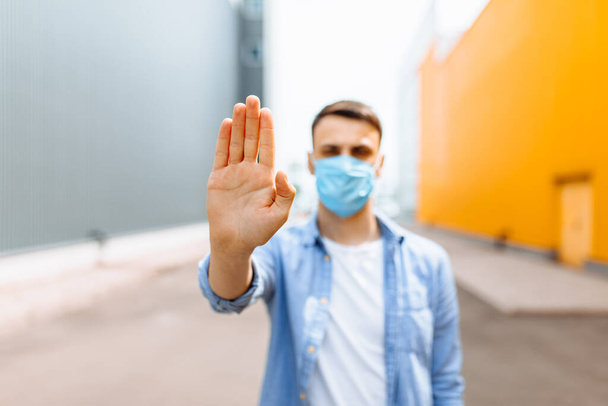 A young man in a protective medical mask makes a stopping hand gesture while standing in the city. Quarantine, epidemic, coronavirus - Foto, Imagem