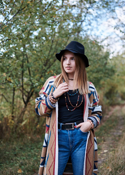 Three-quarter portrait of pretty blond woman in autumn park forest woods. Young girl, wearing black hat, blue mama jeans and colorful cardigan, posing on natural background. Hippie style. - Photo, Image