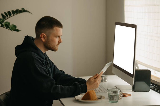 Fernarbeit online. Ein Programmierer arbeitet aus der Ferne mit einem All-in-One-Computer. Ein Kollege hält von zu Hause aus einen Tablet-Computer in der Hand. - Foto, Bild