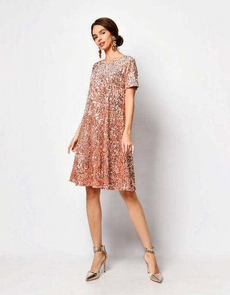 Glamour mladá fena se stylovým účesem a tlustými náušnicemi předvádějící třpytivě krátké korálové růžové šaty a stříbrné boty izolované na bílém - Fotografie, Obrázek