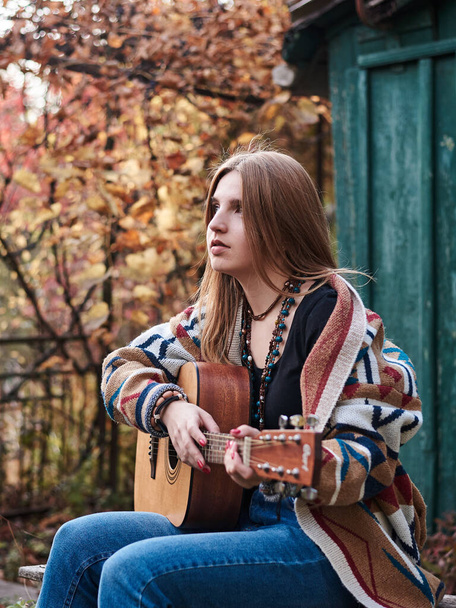 Young blond woman, wearing colorful cardigan, holding acoustic guitar, sitting on a bench in front of old green wooden hut in the woods in autumn. Three-quarter portrait of hippie musician in village - Foto, Imagen