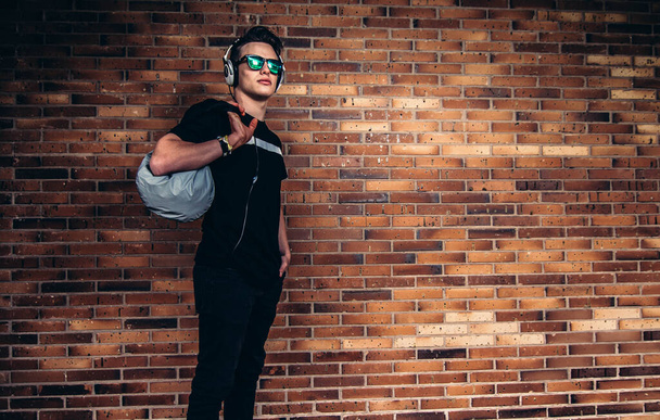 Jovem homem bonito vestindo óculos de sol e segurando saco esportivo contra parede de tijolo
. - Foto, Imagem