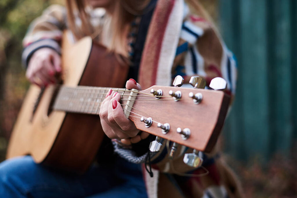 Young woman, wearing colorful cardigan, playing acoustic guitar, Close-up picture of guitar fingerboard, held in hands with bracelets of hippie musician. Playing guitar. - Фото, изображение