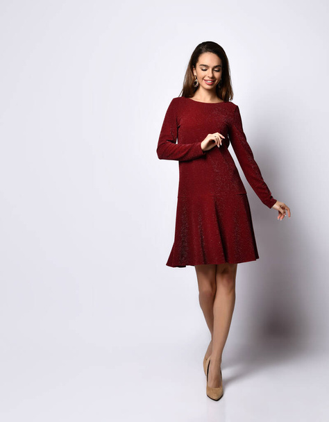 smiling young female in a rich red long-sleeved flying gown and nude shoes standing cross-legged putting hand on hip, isolated on white - Foto, afbeelding