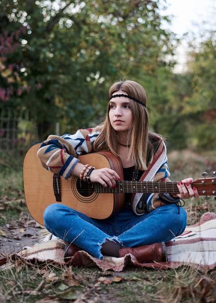 Young blond woman, wearing colorful cardigan and blue jeans, holding acoustic guitar, sitting on blanket in countryside in autumn. Hippie musician, playing guitar outside in the village park - Foto, Imagen