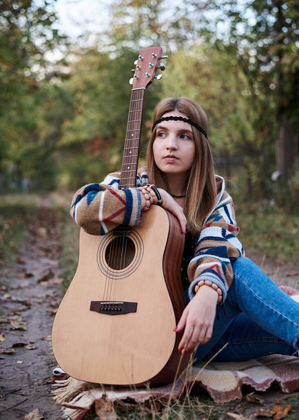 Young blond woman with bracelets on her hands wearing colorful cardigan and blue jeans, holding embracing acoustic guitar, sitting on ground in countryside. Hippie musician with guitar outside in park - Φωτογραφία, εικόνα