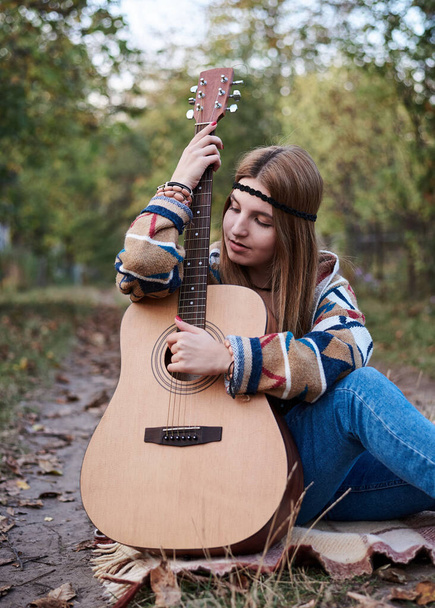 Young blond woman with bracelets on her hands wearing colorful cardigan and blue jeans, holding embracing acoustic guitar, sitting on ground in countryside. Hippie musician with guitar outside in park - Foto, Imagen