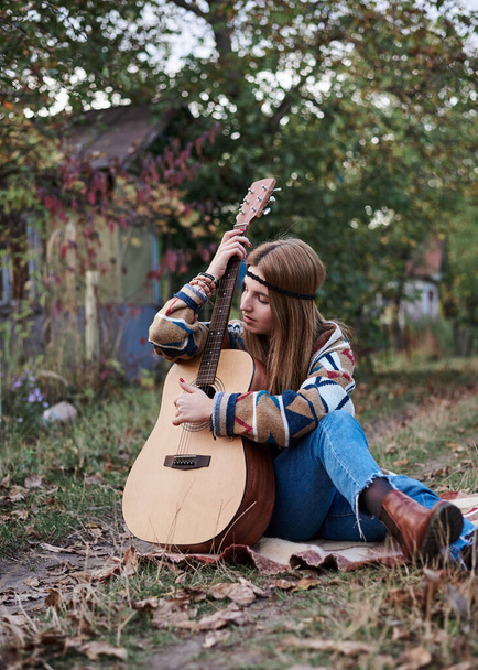 Young blond woman with bracelets on her hands wearing colorful cardigan and blue jeans, holding embracing acoustic guitar, sitting on ground in countryside. Hippie musician with guitar outside in park - Photo, Image