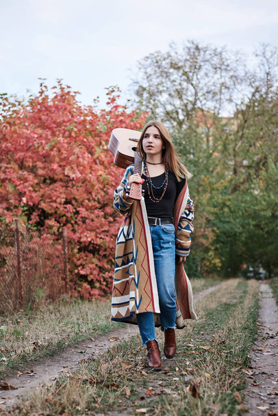Young blond woman, wearing colorful cardigan and blue jeans, holding acoustic guitar on her shoulder, walking in park woods forest in autumn. Full-length portrait of hippie musician with guitar. - Foto, Bild