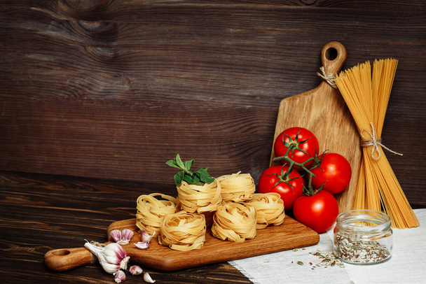 Raw spaghetti and fettuccine with ingredients for cooking pasta on wooden table with blank of wooden kitchen board. Rustic style. Copy space - Photo, Image