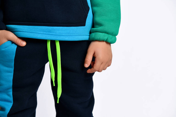 Little male in colorful tracksuit. He has put one hand in pocket, posing isolated on white studio background. Fashion, advertising - 写真・画像