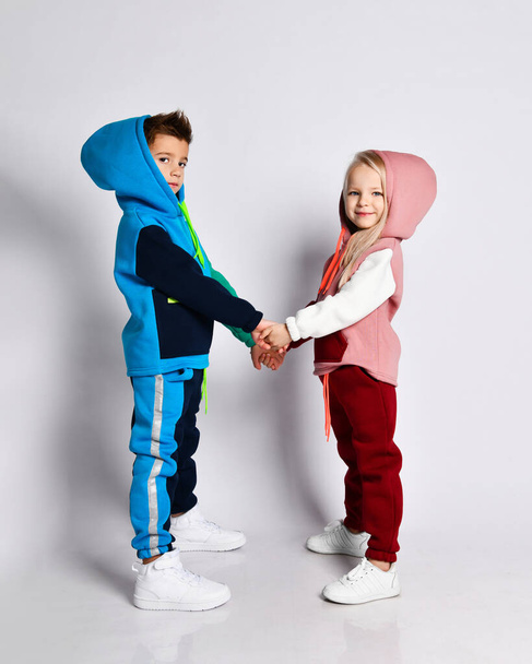 Little kids, brother and sister, in hoods, colorful tracksuits and sneakers. They holding hands, posing sideways isolated on white - Photo, Image