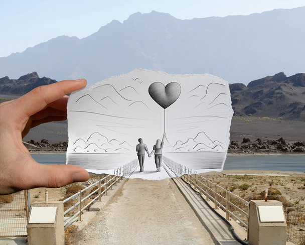 Gemengde mediabeelden van een hand-held stuk papier met een schets op het beeld van een wandelend paar met de vrouw met een grote hartvormige ballon, met een rotsachtig landschap op de foto-achtergrond - Foto, afbeelding