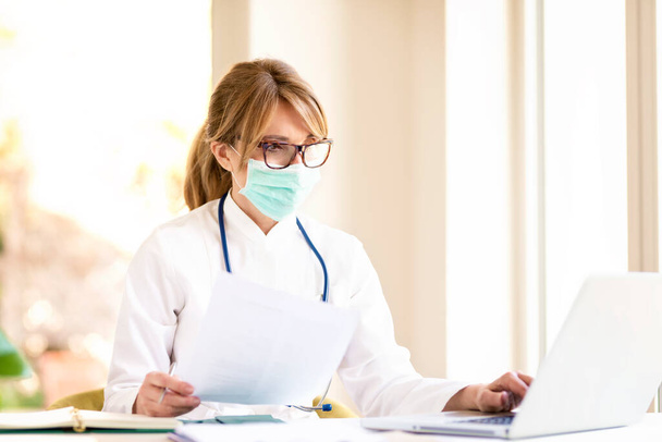 Shot of overworked female doctor wearing face mask for prevention while sitting at desk and working on laptop in the hospital. - Foto, immagini