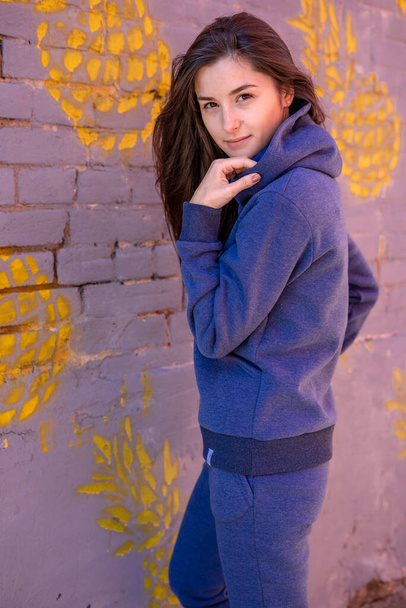 Stylish, beautiful, smiling, young woman wearing a warm, sporty suit in blue and with a hood, poses for photos in the background of city streets. Street home style clothing for women and girls. - Photo, image