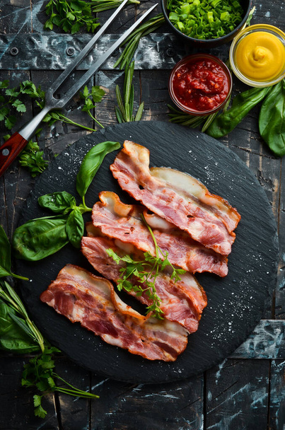 Fried bacon slices with basil on a black stone plate. Top view. Free copy space. - Photo, image