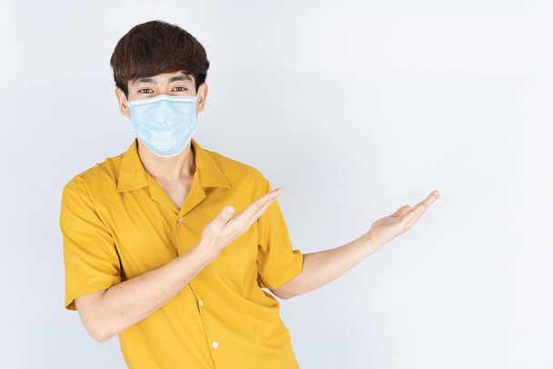 Ασιάτης όμορφος χαρούμενος hipster άνδρας φορώντας μια μάσκα προσώπου ιατρικής προστασίας για την προστασία του coronavirus, φίλτρο σκόνη PM 2.5 και κρύο και το δάχτυλό της δείχνει έξω στο λευκό φόντο - Φωτογραφία, εικόνα