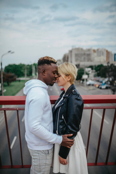 Young interracial couple stands on bridge and hugs against background of road and city. Concept of love relationships and unity between different human races. - Photo, Image