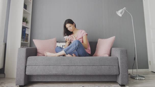 Nuori söpö nainen käyttää puhelinta istuessaan sohvalla kotona. - Materiaali, video