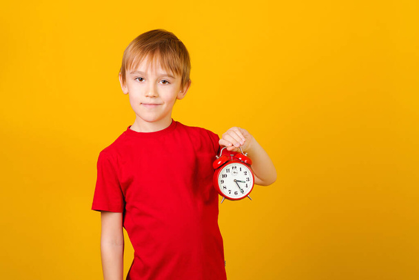 Cute boy holding red alarm clock. Time management concept. Schoolboy with retro clock isolated over yellow background. Time concept. Mock up, copy space. Vintage clock in a hand. - Fotó, kép