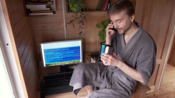 freelance developer in bathrobe remote working from home office in pandemic quarantine, talking on smartphone and smile, coughs or sneezes, concept of self-isolated lifestyle workspace or workplace - Footage, Video