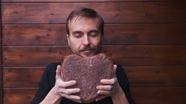 bearded baker with love holds, sniffs and enjoys aroma of handmade freshly baked vegan rye whole grain sourdough bread in shape of heart, smiles and holds out loaf, valentine gift - Footage, Video