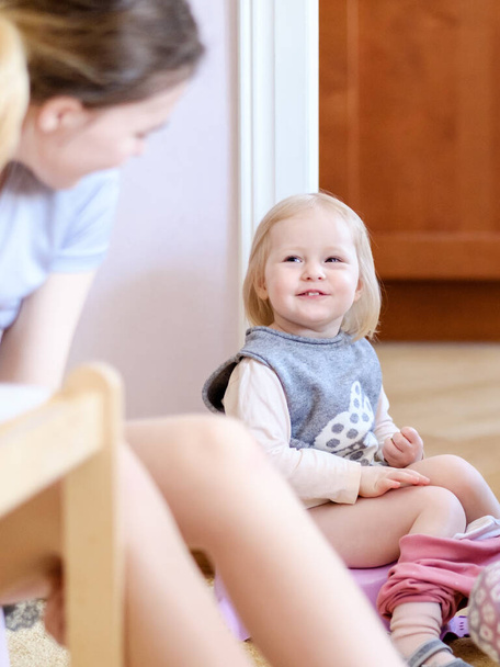 Little blond girl child sits on a baby potty and mischievously looks at her older sister. A happy child laughs, freaks out and has fun. The child pees and poops. - 写真・画像