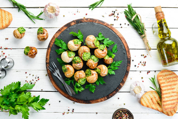 Baked snails with garlic butter and fresh herbs on a black plate on a white wooden background. Top view. Free space for your text. - Zdjęcie, obraz