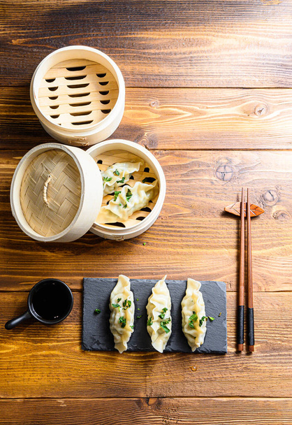 Asian dumplings on stone slate and in wooden steamer over traditional wood table with soy sauce and chopsticks and fresh herbs top view space for text. - Foto, immagini