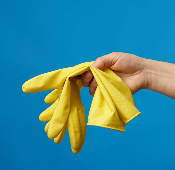 női kéz tartja sárga gumikesztyű tisztítására, kék háttér, védőruházat a ház tisztítására - Fotó, kép