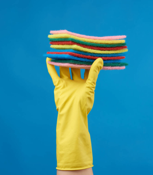 掃除用の黄色いゴム手袋を手に身体の一部を持ち上げ青い背景にキッチンスポンジの山を持ち上げ - 写真・画像