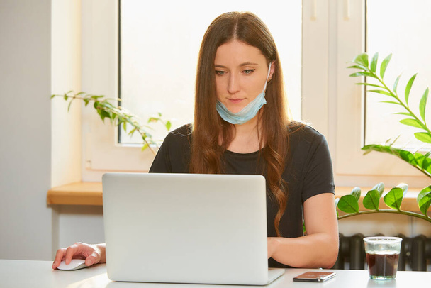 Een vrouw in het verwijderde medische gezichtsmasker werkt op afstand op haar laptop tijdens de quarantaine om het verspreide coronavirus te voorkomen. Een zakenvrouw belt vanuit huis tijdens de pandemie van COVID-19. - Foto, afbeelding