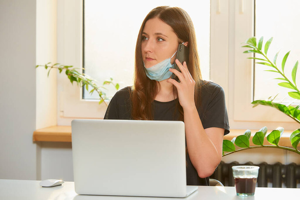 A woman in the removed medical face mask working remotely on her laptop during the quarantine to avoid the spread coronavirus. A businesswoman calling from home during the pandemic of COVID-19. - Photo, image