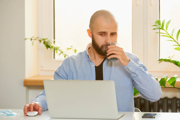 Человек, пьющий кофе, работая дистанционно над своим ноутбуком во время карантина, чтобы избежать распространения коронавируса. Лысый бизнесмен с бородой, работающий из дома во время пандемии COVID-19
. - Фото, изображение