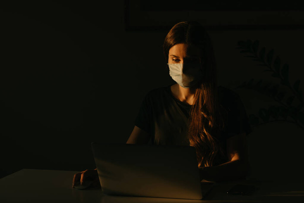 Una donna in maschera medica che studia a distanza sul suo portatile durante la quarantena per evitare il coronavirus diffuso. Una ragazza che lavora da casa la sera durante la pandemia di COVID-19
. - Foto, immagini