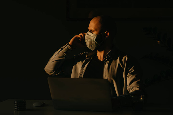 Un uomo con una maschera medica che lavora a distanza sul suo portatile durante la quarantena per evitare il coronavirus diffuso. Un uomo d'affari che chiama da casa la sera durante la pandemia del COVID-19
. - Foto, immagini