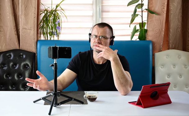 Evde akıllı telefon ve tabletle video konferansı düzenlerken konuşan kulaklı ve gözlüklü yetişkin bir adamın portresi. Siyah tişörtlü bir model kameranın tam karşısında oturuyor.. - Fotoğraf, Görsel