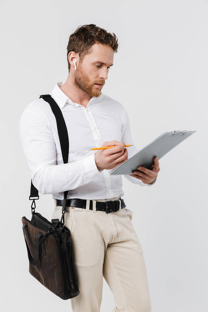 Image of focused man in wireless earphones holding clipboard and writing isolated over white background - Zdjęcie, obraz