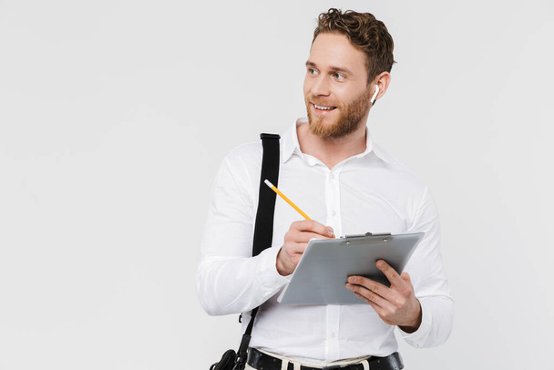 Image of smiling man in wireless earphones holding clipboard and writing isolated over white background - Фото, зображення