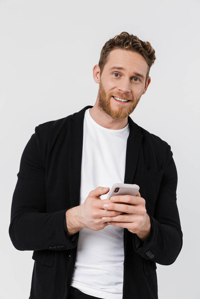 Bild von gut aussehenden fröhlichen Mann in Jacke lächelnd und mit Smartphone isoliert über weißem Hintergrund - Foto, Bild