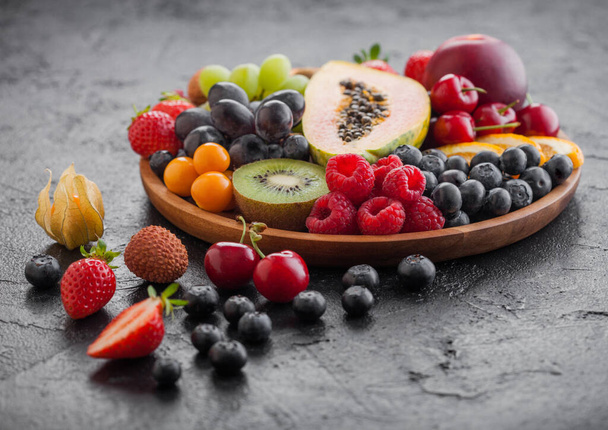 Fresh raw organic summer berries and exotic fruits in round wooden plate on black kitchen background. Papaya, grapes, nectarine, orange, raspberry, kiwi, strawberry, lychees, cherry. Top view - Fotografie, Obrázek