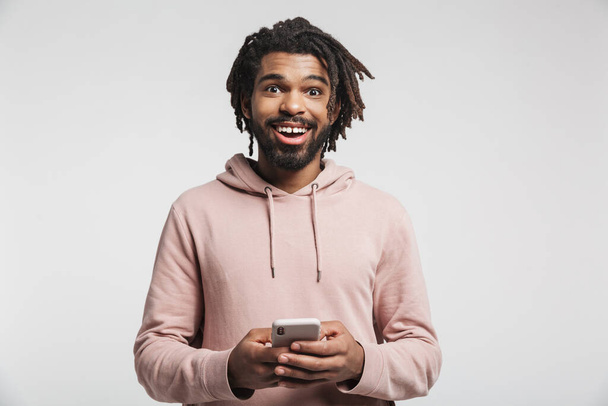 Portrait of a young african man wearing hoodie standing isolated over white background, using mobile phone - Foto, Imagem