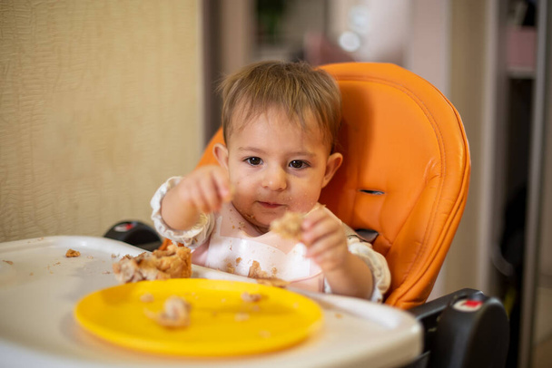 Um bebê bonito em um assento infantil laranja com uma mesa, segura um pedaço de bolo e olha para ele. há migalhas e um prato laranja na mesa. close-up, vista frontal, foco suave, fundo desfocado
 - Foto, Imagem