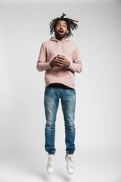 Full length portrait of a young african man wearing hoodie jumping isolated over white background, holding mobile phone - Φωτογραφία, εικόνα