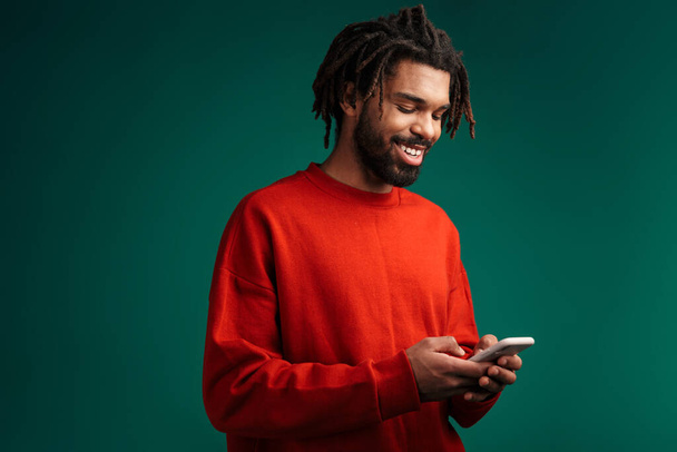 Portret van een jonge Afrikaanse man die een pullover draagt die geïsoleerd staat boven groene achtergrond, met behulp van een mobiele telefoon - Foto, afbeelding