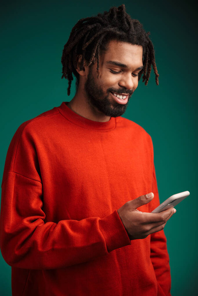 Porträt eines jungen afrikanischen Mannes mit Pullover, der isoliert auf grünem Hintergrund steht und Mobiltelefone benutzt - Foto, Bild